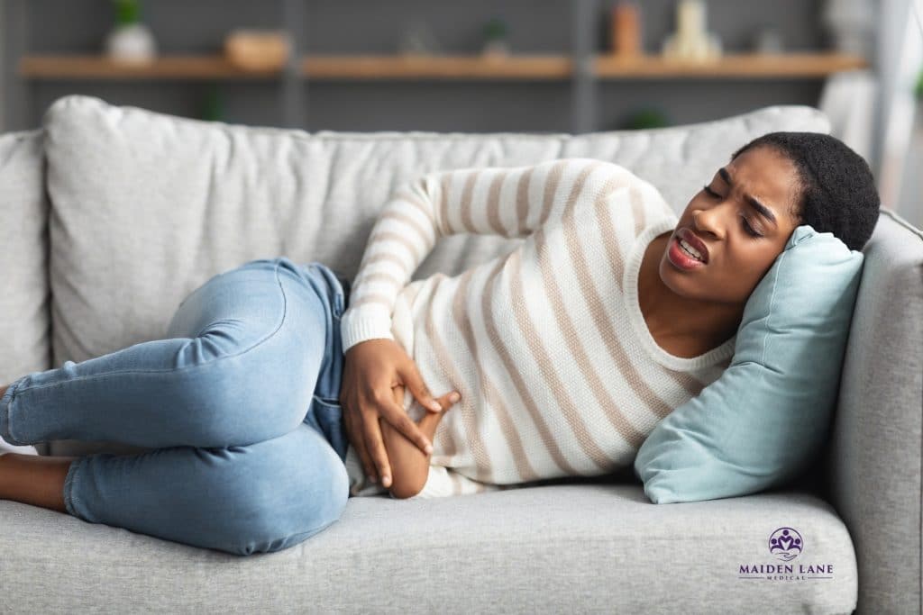 Young African American woman laying on her couch holding her stomach. Maiden Lane Medical | New York, NY